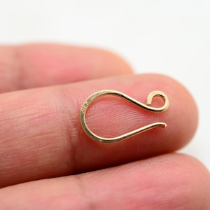 May include: A small, gold-coloured jewellery clasp with a curved design. The clasp has a hook shape with a looped end and a curved bottom. The jewellery clasp is held between two fingers against a white background.