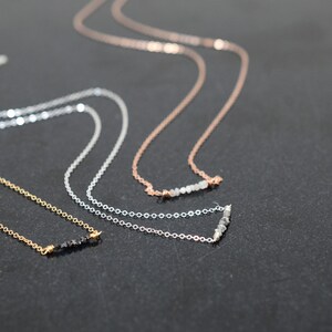 Puede incluir: Tres delicados collares con cadenas de diferentes colores y peque&ntilde;os detalles de piedras preciosas. Los collares est&aacute;n dispuestos sobre una superficie oscura.