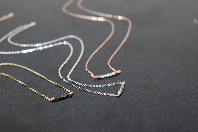 Puede incluir: Tres delicados collares con cadenas de diferentes colores y peque&ntilde;os detalles de piedras preciosas. Los collares est&aacute;n dispuestos sobre una superficie oscura.