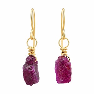 May include: A pair of gold-colored earrings featuring raw, deep pink ruby gemstones. The ruby stones are wire-wrapped and suspended from gold-colored ear wires. The earrings are set against a white background.