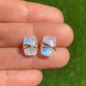 May include: A pair of earrings featuring iridescent moonstone stones wrapped in gold wire. The stones are rectangular in shape and have a white, blue, and iridescent sheen.