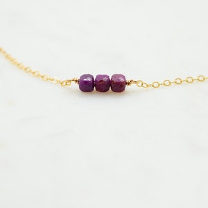 May include: A delicate gold chain necklace with three small, square-shaped purple gemstones.