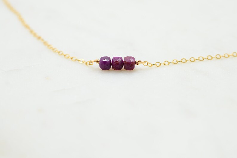 May include: A delicate gold chain necklace with three small, square-shaped purple gemstones.
