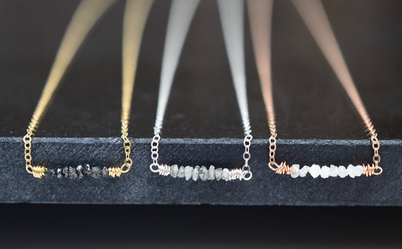 Puede incluir: Tres delicados collares con una fila de peque&ntilde;os diamantes en bruto engastados en un dise&ntilde;o simple de alambre. Los collares se muestran sobre una superficie negra y est&aacute;n disponibles en oro, plata y oro rosa.