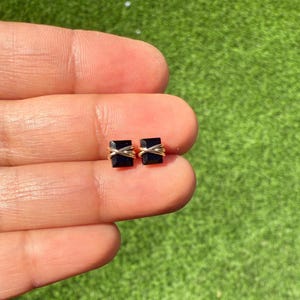May include: A pair of square black gemstone stud earrings with gold-colored wire detailing. The earrings are displayed on a hand against a green background. The earrings are small and minimalist.