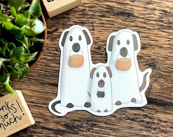 May include: Three white ghost dogs with black eyes and noses. The two larger dogs are holding orange pumpkins and the smaller dog is holding a black cauldron. The dogs are on a wood background.