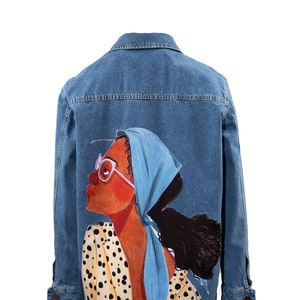 Puede incluir: Una chaqueta vaquera azul con una ilustración colorida de una mujer con un pañuelo azul y gafas en la espalda.