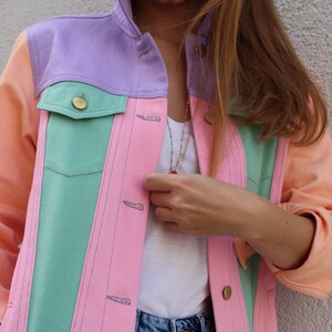 Puede incluir: Una chaqueta vaquera de color rosa pastel, verde y morado con una camiseta blanca y unos vaqueros azules debajo.
