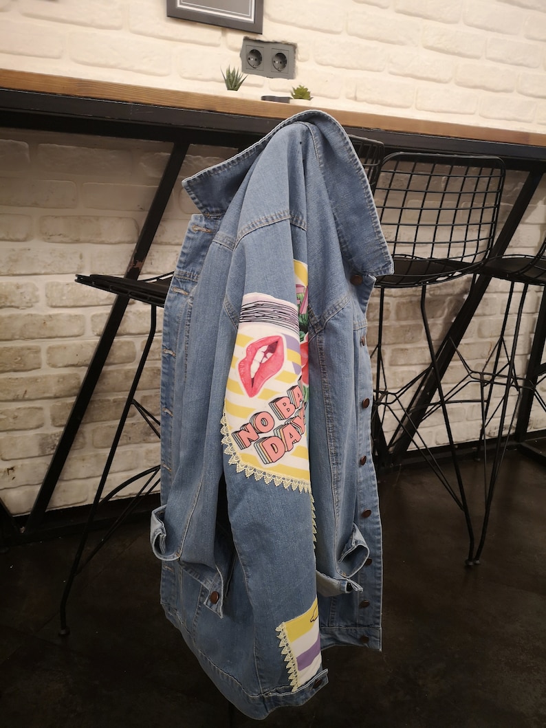May include: A light blue denim jacket with a colorful graphic print on the left sleeve. The print features a red mouth, yellow stripes, and the text "NO BAD DAYS".