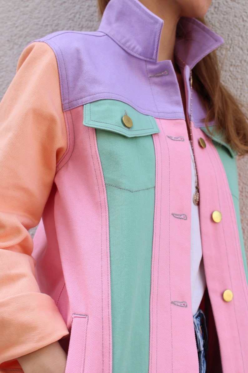 Puede incluir: Una chaqueta vaquera de color rosa pastel, verde y morado con cierre de botones. La chaqueta tiene un bolsillo de parche en el pecho izquierdo y dos bolsillos de solapa en la cintura.