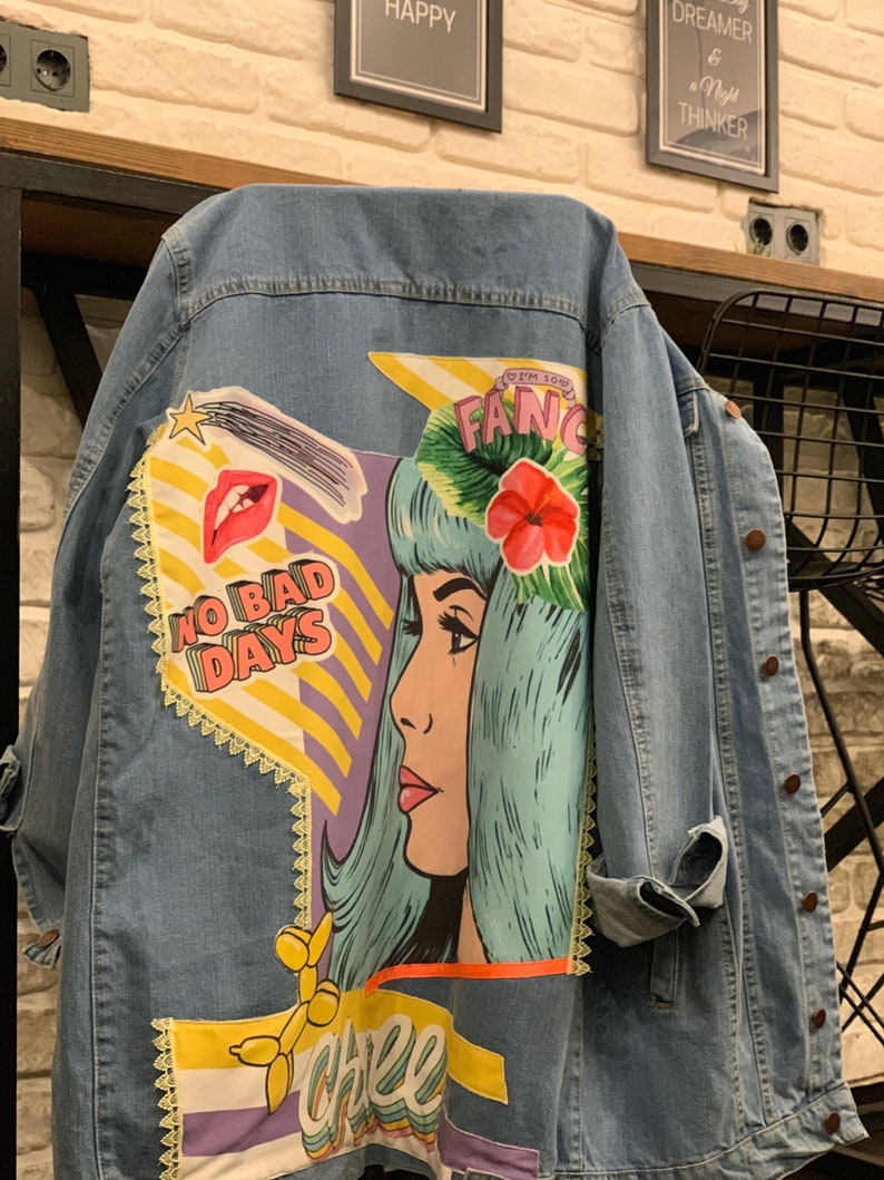 May include: A blue denim jacket with colorful patches and embroidery. The patches feature a cartoon woman with blue hair, a red flower, and the words "No Bad Days" and "Cheer".