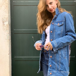 May include: A woman wearing a long, blue denim jacket over a white t-shirt and blue jeans. She is holding a pair of sunglasses in her hand.