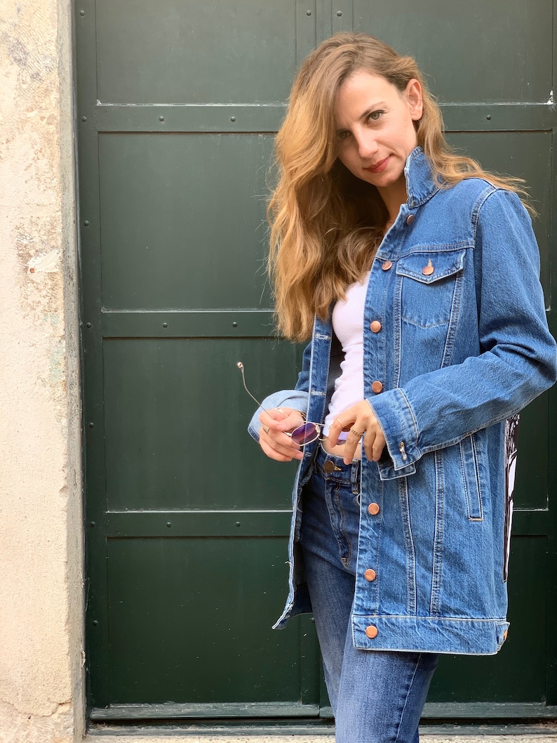 May include: A woman wearing a long, blue denim jacket over a white t-shirt and blue jeans. She is holding a pair of sunglasses in her hand.