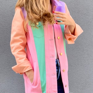 Puede incluir: Una chaqueta vaquera rosa con un dise&ntilde;o de bloques de color verde, morado y naranja. La chaqueta tiene cierre de botones y bolsillos.
