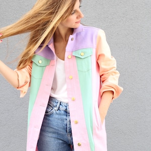 Puede incluir: Una chaqueta vaquera de color pastel con un esquema de color rosa claro, verde claro y lavanda. La chaqueta tiene botones dorados y un estilo cl&aacute;sico de chaqueta vaquera.