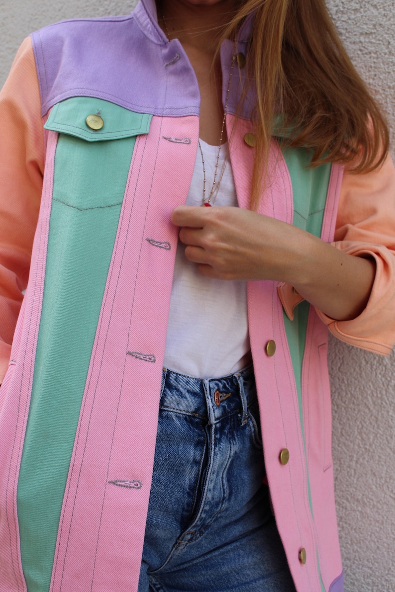 Puede incluir: Una chaqueta vaquera de color rosa pastel, verde y morado con una camiseta blanca y unos vaqueros azules debajo.