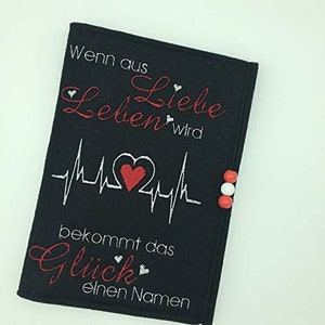 Könnte beinhalten: Schwarze Stoff-Notizbuchhülle mit weißer und roter Stickerei. Der Text lautet "Wenn aus Liebe Leben wird bekommt das Glück einen Namen".