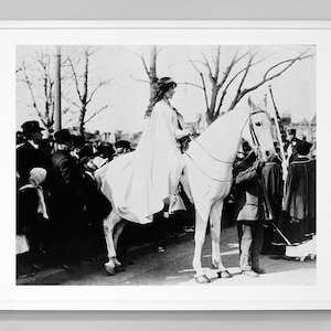 May include: A woman in a white dress and cape rides a white horse through a crowd of people. The woman is wearing a crown and holding a bouquet of flowers.