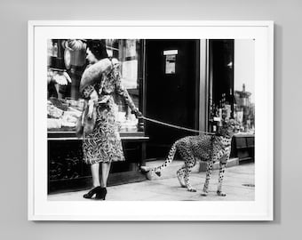 May include: A woman in a patterned dress walks down a city street while holding a leash attached to a cheetah.