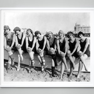 May include: A black and white photograph of eight women wearing swimsuits and hats, sitting on a wooden bench on a beach. They are all smiling and looking at the camera.