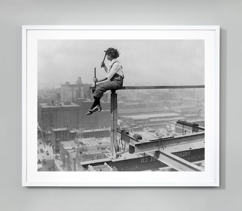 Pu&ograve; includere: Una fotografia in bianco e nero di una donna seduta su una trave in alto sopra una citt&agrave;. Indossa una camicia a maniche lunghe e pantaloni. La citt&agrave; &egrave; sullo sfondo ed &egrave; composta da molti edifici.