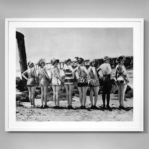Könnte beinhalten: Schwarzweißfoto von acht Frauen in Vintage-Badeanzügen, die an einem Strand vor einem Pier stehen.