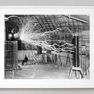 May include: A black and white photograph of a man sitting in a chair in a large room with a large electrical coil in the background. The coil is producing a large electrical arc, which is visible in the photograph. The man is wearing a suit and tie, and he is looking at the electrical arc.