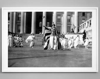 May include: A black and white photograph of a woman in a costume holding a flag in front of a building. She is surrounded by other people in white robes.