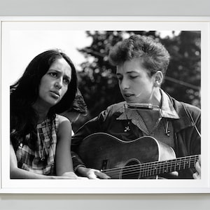 May include: A black and white photograph of a man and a woman. The man is playing a guitar and harmonica. The woman is looking at the camera.