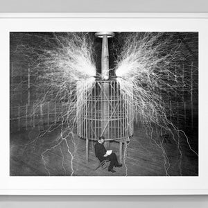 May include: A black and white photograph of a man sitting in a chair in front of a large electrical transformer. The transformer is emitting sparks of electricity.