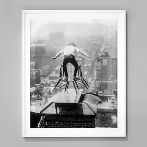 May include: A black and white photograph of a person balancing on two chairs on top of a building. The person is wearing a long-sleeved shirt and trousers. The city skyline is visible in the background.