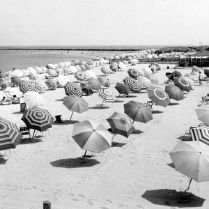 May include: A black and white photo of a beach with many striped umbrellas and chairs. The umbrellas are arranged in rows on the sand. There are people in the background.