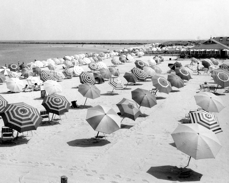 May include: A black and white photo of a beach with many striped umbrellas and chairs. The umbrellas are arranged in rows on the sand. There are people in the background.