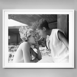 May include: A black and white photograph of a couple sharing a cigarette. The woman is on the left, wearing a white tank top and looking at the man. The man is on the right, wearing a white tank top and looking at the woman. They are both smiling.