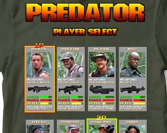 Könnte beinhalten: Ein grünes T-Shirt mit einer Retro-Videospielgrafik der Predator-Filmfiguren. Die Grafik zeigt den Text "Predator Player Select" und zeigt die verschiedenen Charaktere aus dem Film mit ihren Waffen, ihrer Geschwindigkeit, ihrer Kraft und ihren Fähigkeiten.