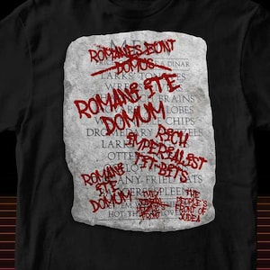 Puede incluir: Camiseta negra con un diseño gráfico de una tableta de piedra con texto rojo, incluyendo "ROMANES BUNT DOMUS...". Algunas palabras están tachadas. La camiseta es de cuello redondo.
