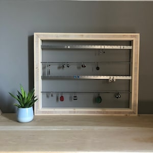 Op de afbeelding: Een houten oorbellenhouder met meerdere rijen metalen haken om oorbellen aan te hangen. De houder is aan een muur gemonteerd en heeft een lichte houtkleur.