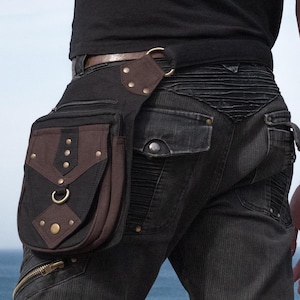 May include: Black and brown hip bag with a decorative design. The bag has a flap with a brass-coloured ring and studs. The bag is attached to a belt loop with a leather strap. The person is wearing black jeans.