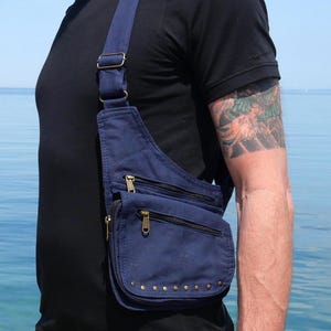 Puede incluir: Un bolso bandolera azul marino con múltiples compartimentos con cremallera y una solapa con tachuelas. El bolso tiene una correa ajustable y herrajes de color latón. El bolso está hecho de lona duradera.