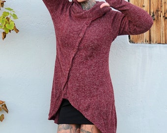 Op de afbeelding: Een vrouw draagt een bordeauxrode hoodie met een enkele knoopsluiting. De trui heeft een lange, asymmetrische zoom. Ze draagt een zwart hoofdband en heeft rood haar.