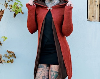 Op de afbeelding: Een vrouw draagt een lange, roestkleurige capuchontrui met een zwart onderhemd en een zwarte rok. Ze heeft rood haar en tatoeages op haar benen.