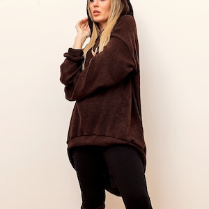 May include: A woman wearing a brown oversized hoodie with a hood pulled up. The hoodie has a long, asymmetrical hemline. She is wearing black leggings and has long blonde hair.