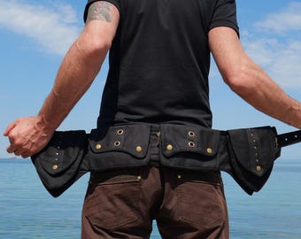 May include: Black utility belt with multiple pockets and compartments. The belt features gold-colored rivets and closures. The belt is worn over brown pants. The background is a blue sky and ocean.