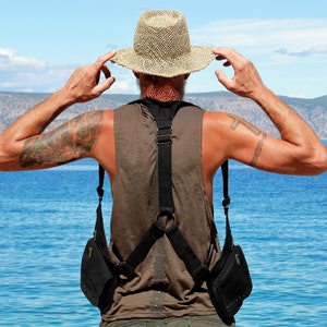 May include: A person wearing a brown sleeveless shirt and a wide-brimmed straw hat, with a black crossbody bag with two pouches. The person is standing with their back to the camera, looking out at a body of water.