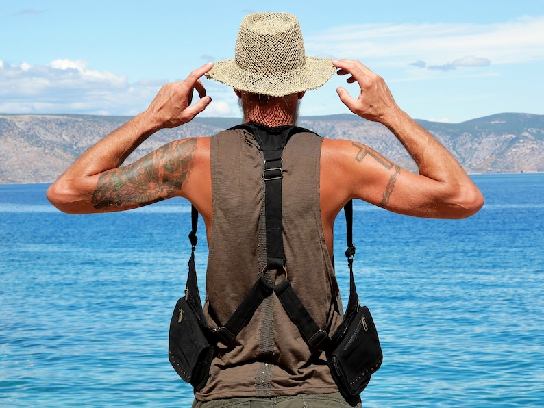 May include: A person wearing a brown sleeveless shirt and a wide-brimmed straw hat, with a black crossbody bag with two pouches. The person is standing with their back to the camera, looking out at a body of water.