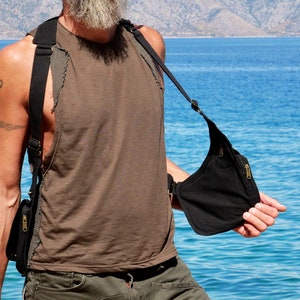 May include: A black canvas crossbody bag with adjustable straps and a zipper closure. The bag is worn by a person standing in front of a body of water.