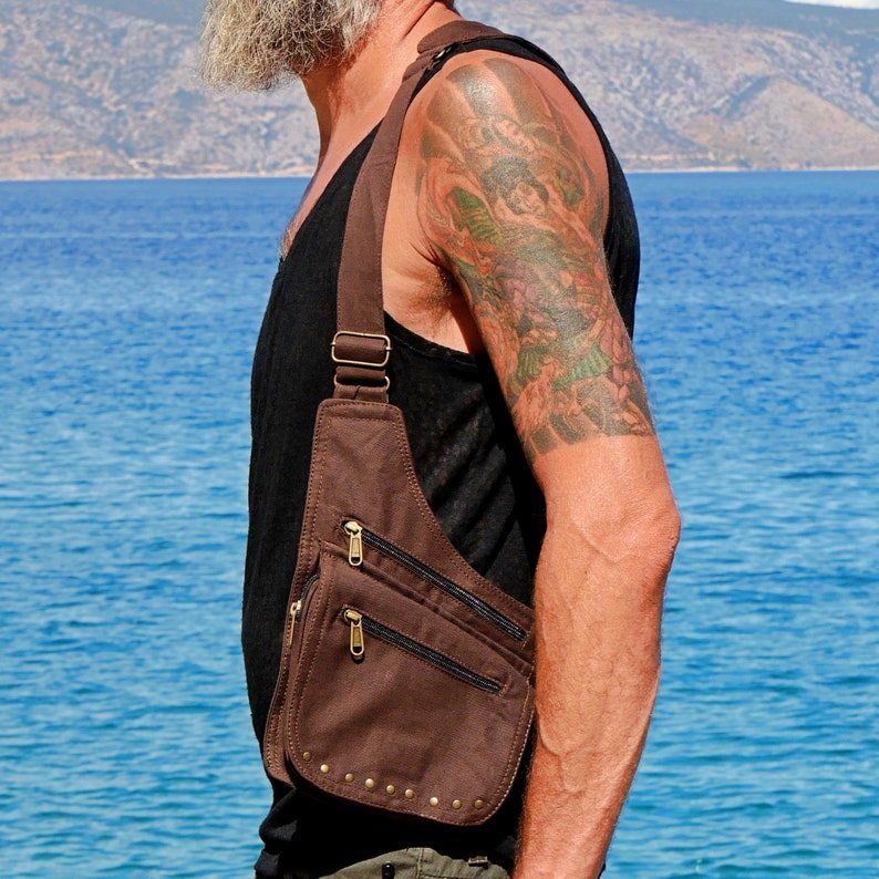 May include: A brown leather crossbody bag with multiple pockets and a zipper closure. The bag is worn by a person with a tattoo on their arm.