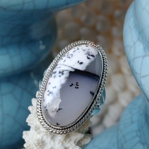 May include: Oval-shaped silver ring featuring a large, polished gemstone. The stone displays a unique pattern of white, gray, and black, with a textured appearance. The ring has a braided silver border, adding a touch of elegance to the jewelry.