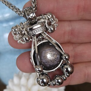 May include: A silver pendant necklace with a dark grey, teardrop-shaped stone set in a decorative metal frame. The pendant has a detailed design with small, round embellishments and hangs from a chain.