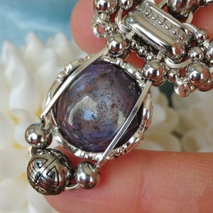May include: A silver pendant with a large, round, purple gemstone. The gemstone is set in a decorative silver frame with a geometric design. The pendant is attached to a chain of small, silver beads and decorative elements.